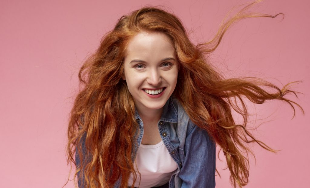 smiling redhead woman red hair flowing leaning forward denim shirt white t-shirt pink background medium porosity hair type what is hair porosity toppik hair blog