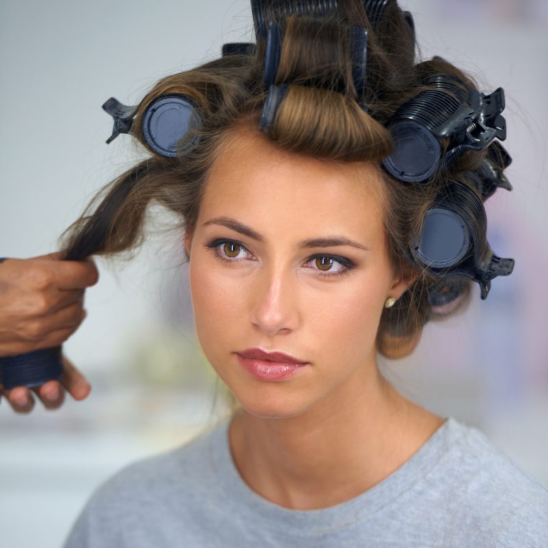 A woman in a gray shirt gets large, volumizing curlers taken out of her hair.