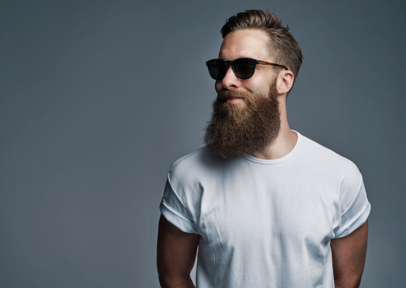 Single bearded young man with serious expression wearing sunglasses and white short sleeve shirt looking over gray background with copy space.