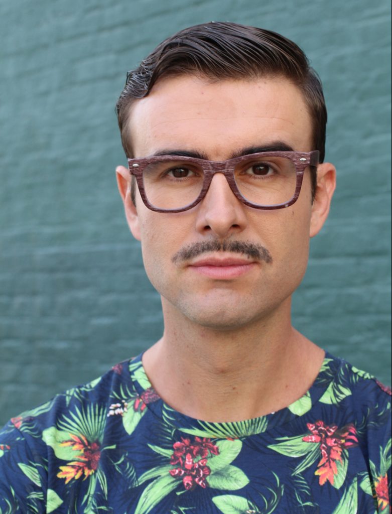 A pencil mustached man wears dark hair glasses and a floral shirt. Perhaps he is participating in movember.