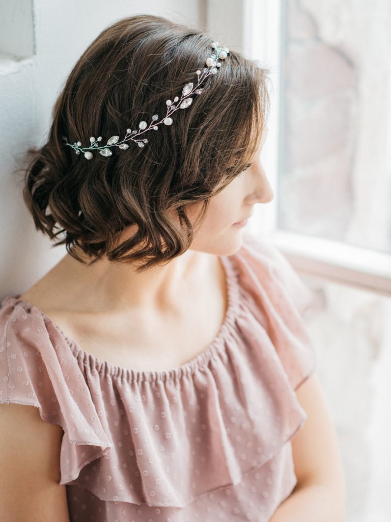 A bridesmaid wearing a crystal headband in her short brown wavy hair gazes out the window.