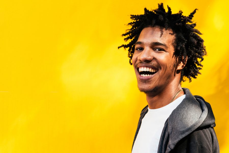 laughing smiling man african american gray hoodie white tee shirt bright yellow background tousled dreads hairstyle 2020 hair trends for men and past styles toppik hair blog