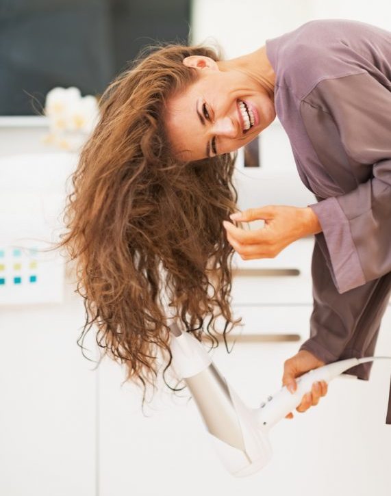 Woman in purple robe laughs while she blow-dries her hair upside down to add volume. How to Add Volume to Thin Hair
