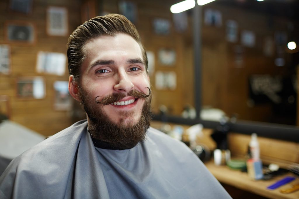 A smiling man sits in a barber shop chair, his handlebar mustache curls up at both corners and his beard is full.