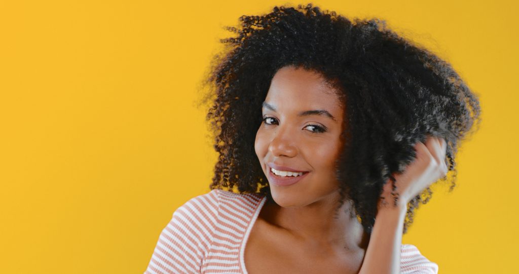 smiling african american woman holding natural hair curly pink white striped shirt yellow background high porosity hair type what is hair porosity toppik hair blog